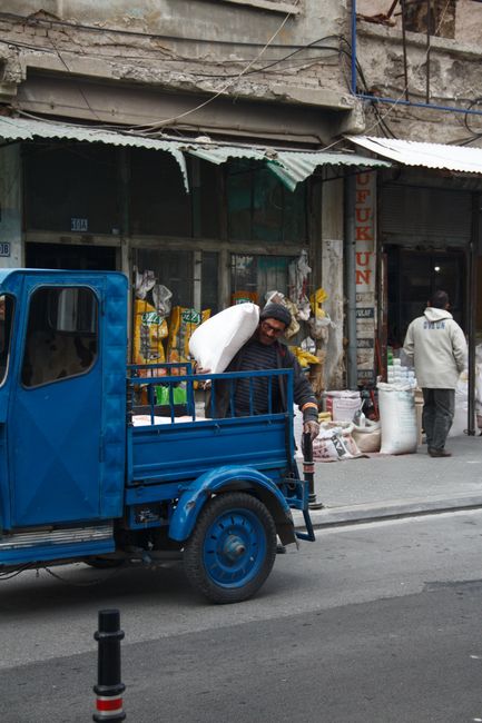 The image depicts a street scene with a small blue truck parked in front of a shop displaying various household goods and packaging materials, including large bags and decorative items. The truck has