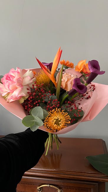 A vibrant bouquet of fresh flowers held by a person wearing black, featuring a mix of pink carnations, purple calla lilies, orange bird of paradise, yellow protea, and red hypericum berries, all wrapp