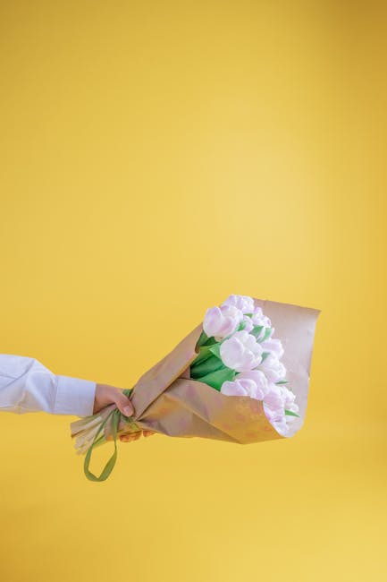 A hand in a white sleeve holds a bouquet of fresh, pale pink tulips wrapped in brown craft paper and tied with a green ribbon, against a bright yellow backdrop. The tulips are vibrant with smooth, rou