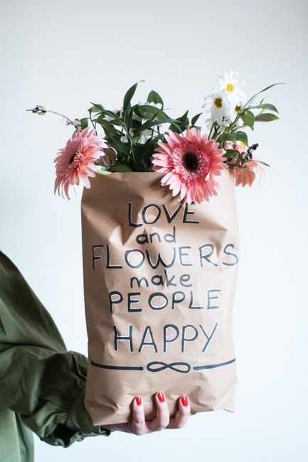 A person wearing a green jacket holds a brown paper bag decorated with a handwritten message in black and blue ink that reads 'LOVE and FLOWERS make PEOPLE HAPPY.' Emerging from the top of the bag is 
