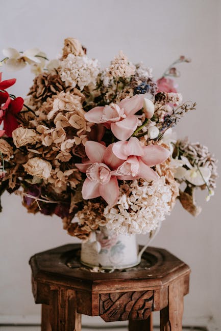 A large bouquet of mixed dried and fresh flowers arranged in a vintage ceramic vase placed on a rustic wooden stool. The arrangement includes soft pink orchids, cream and brown hydrangeas, delicate wh