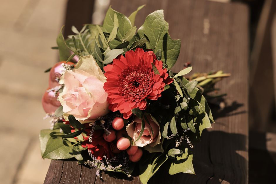A close-up of a floral bouquet featuring a prominent red gerbera daisy with layered petals, surrounded by soft pink roses with delicate, slightly ruffled petals, and accented with small pink berries. 