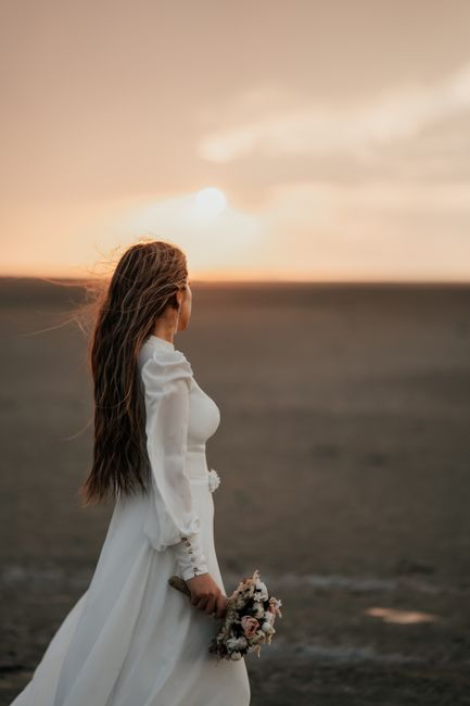 A bride holding a rustic floral bouquet composed of soft pastel roses, peonies, and greenery, wrapped in natural cream-colored paper with a light ribbon, standing on a sandy beach during sunset. She i